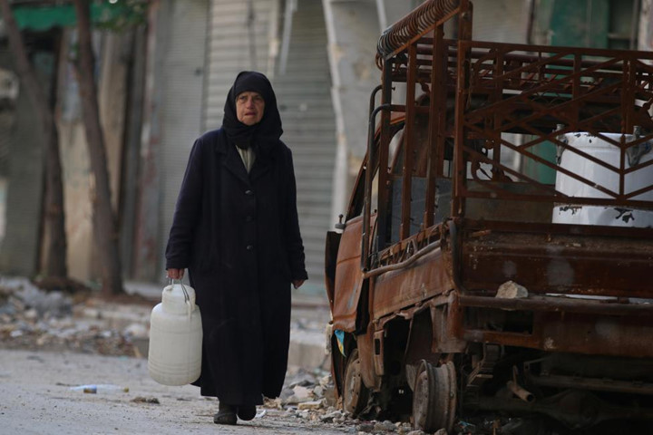 Người phụ nữ luống tuổi mang theo bình đựng nước đứng bên một chiếc xe tải bị cháy rụi ở khu vực Old Aleppo